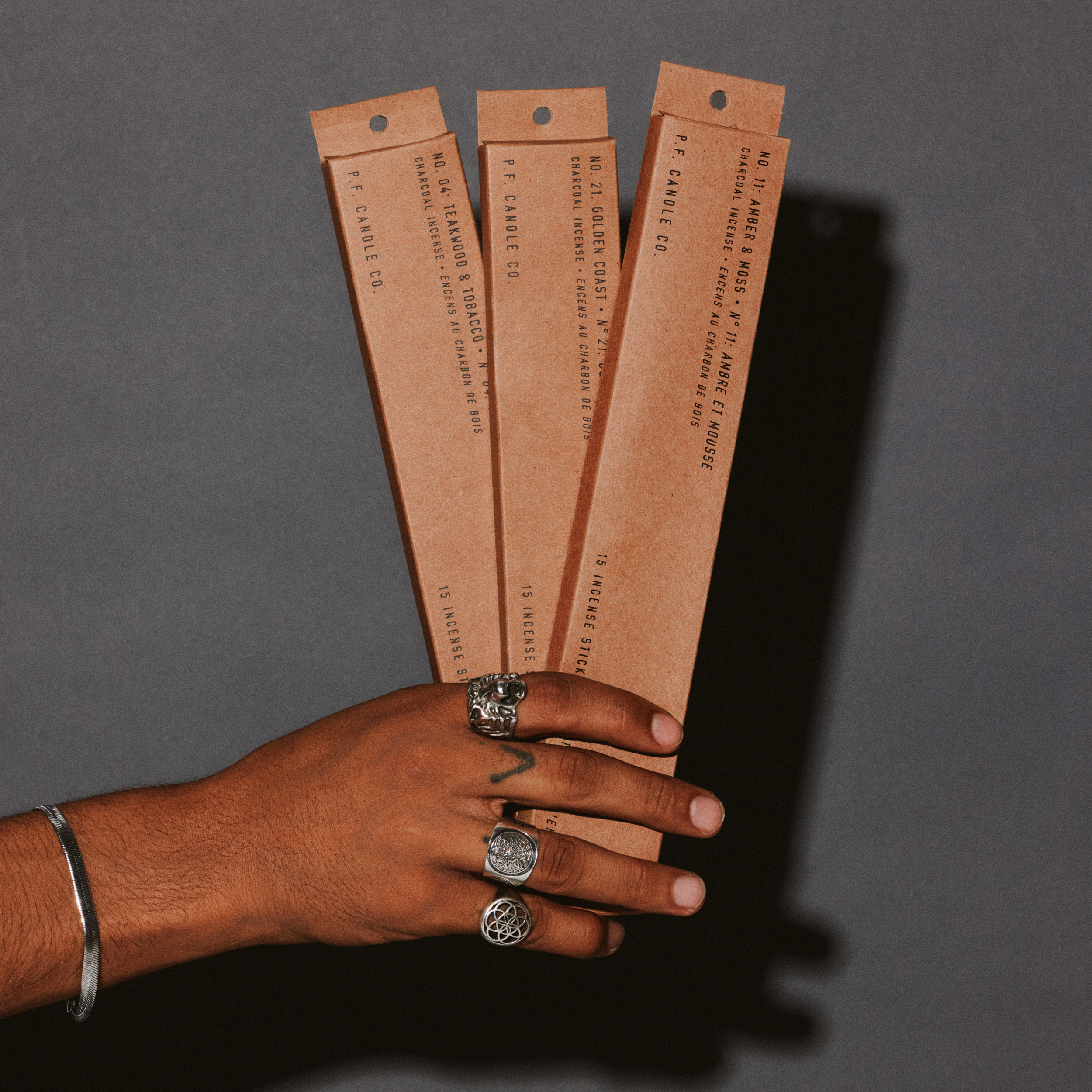 Hand holding three brown kraft paper packages of teakwood & tobacco, golden coast, & amber & moss against a gray background