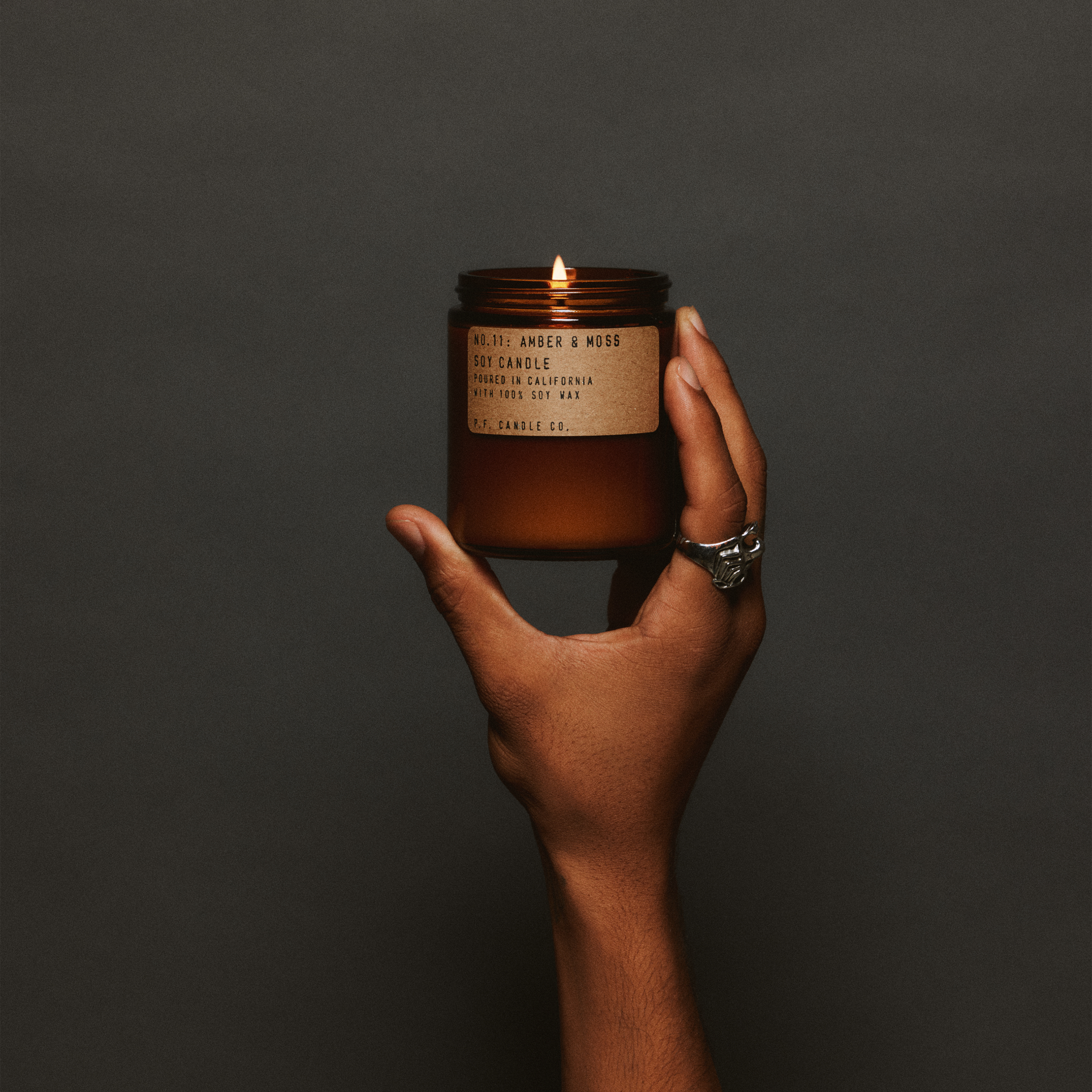 Hand holding a lit amber & moss standard candle with a label against a dark background