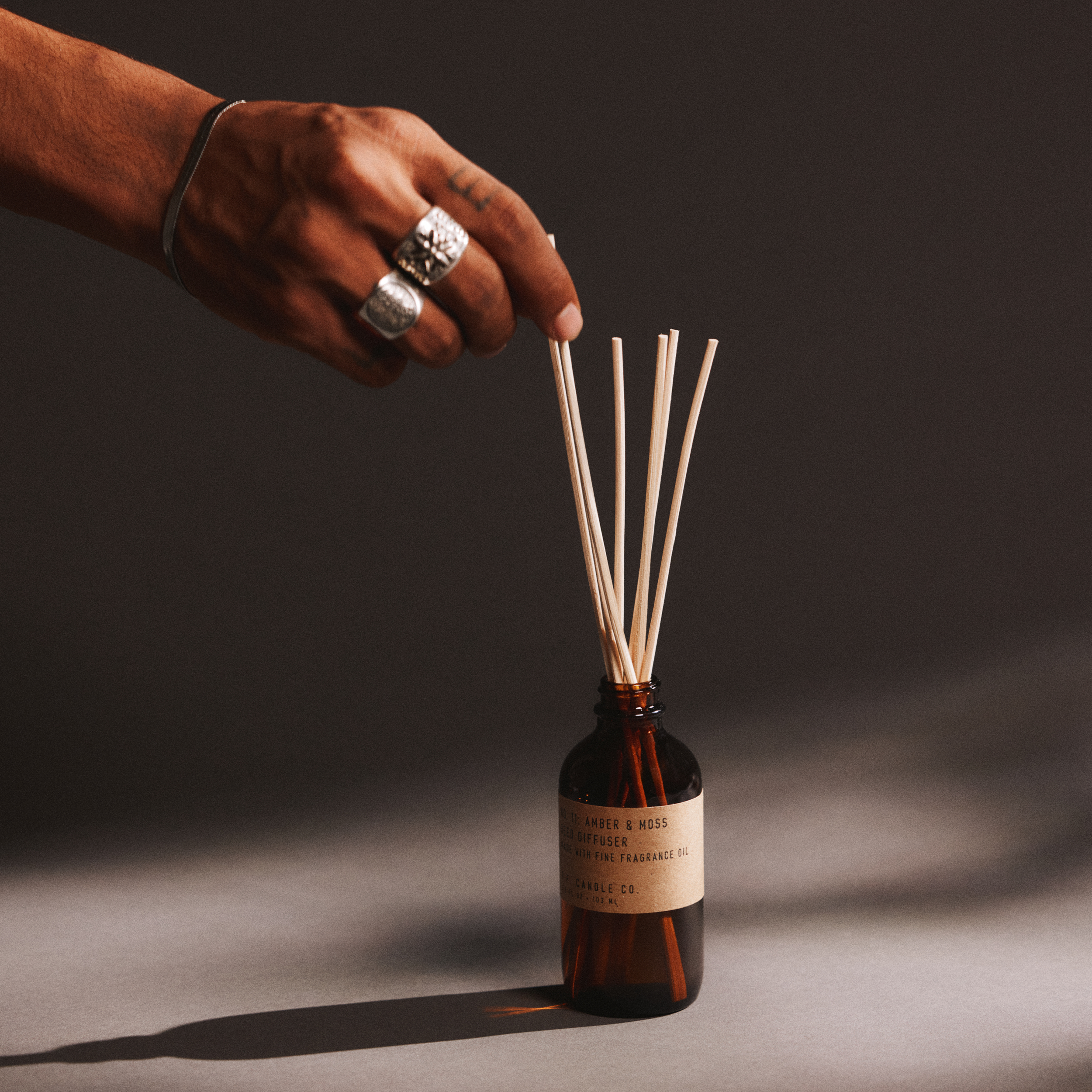 Hand with a ring holding reed diffuser sticks next to a bottle of amber & moss reed diffuser on a dark background