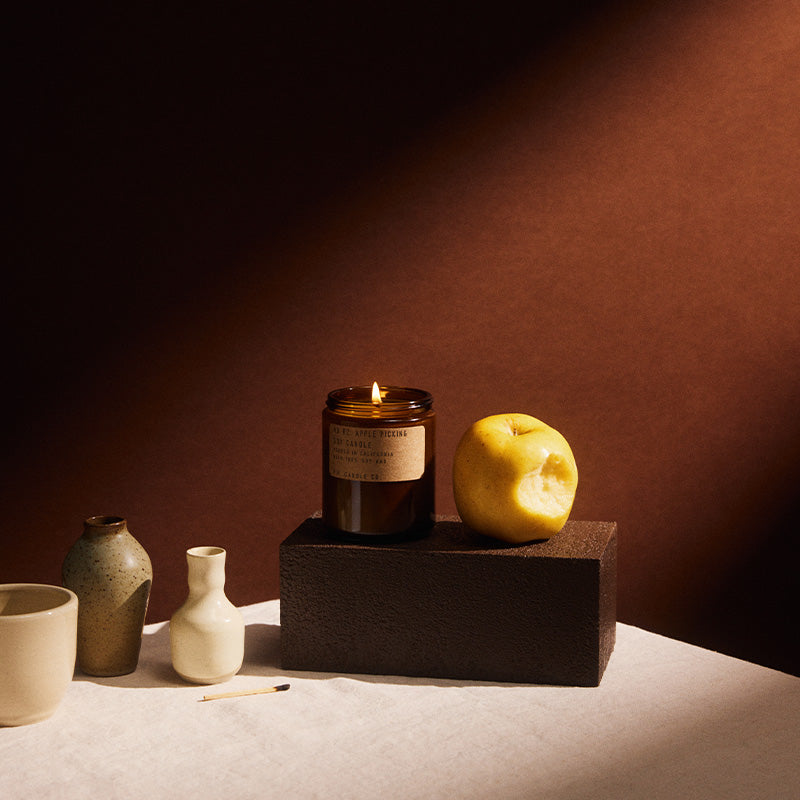 Lit pf candle co no. 2 Apple picking amber jar candle displayed on a pedestal alongside ceramic vases and a cup, beside an apple on a white plate in warm autumn lighting.