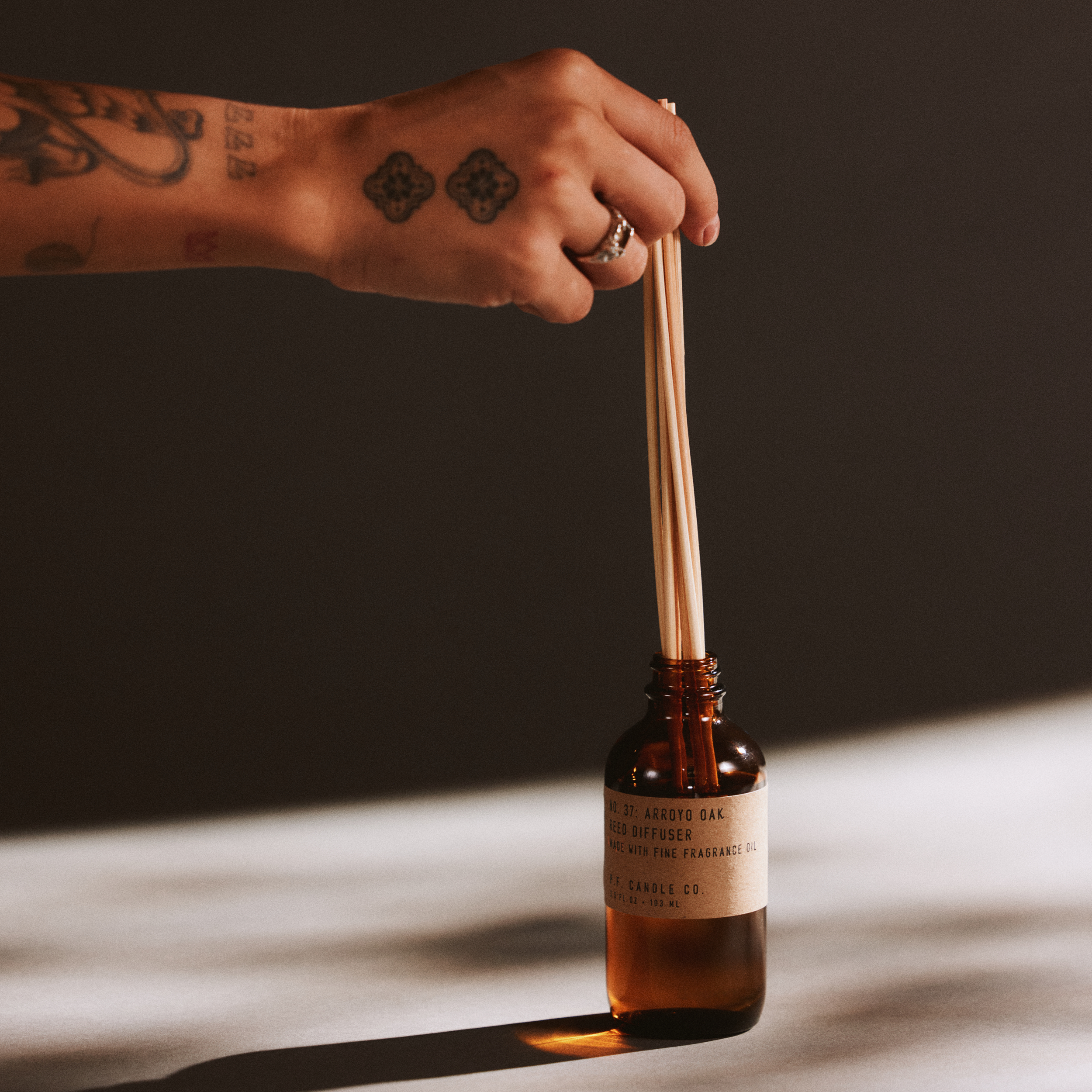 Hand with a ring holding reed diffuser sticks next to a bottle of arroyo oak reed diffuser on a dark background