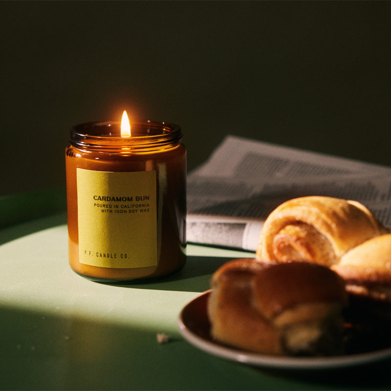 pf candle co Cardamom Bun standard soy candle lit on top of a green table next the a newspaper and cardamom bun
