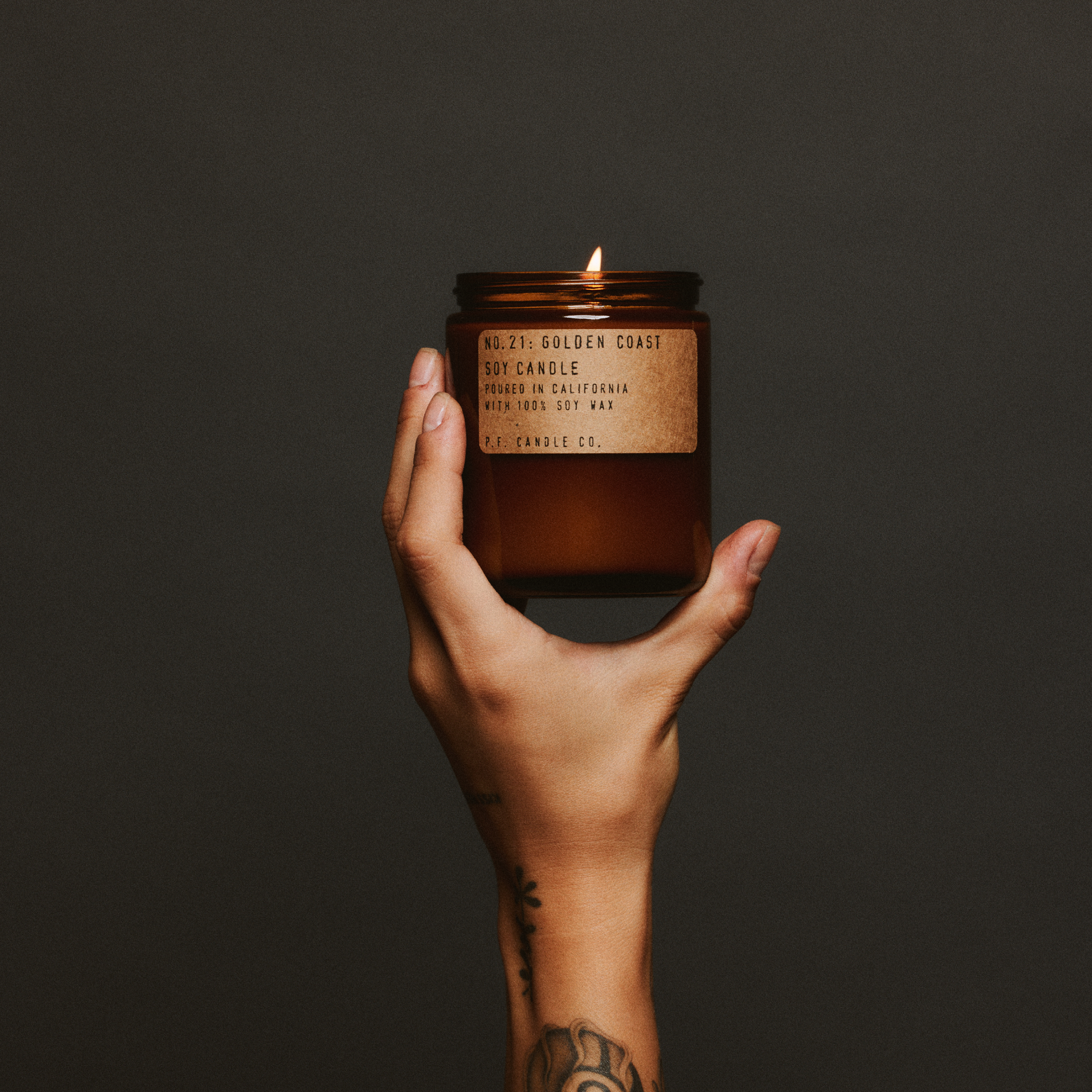 Hand holding a lit golden coast standard candle with a label against a dark background