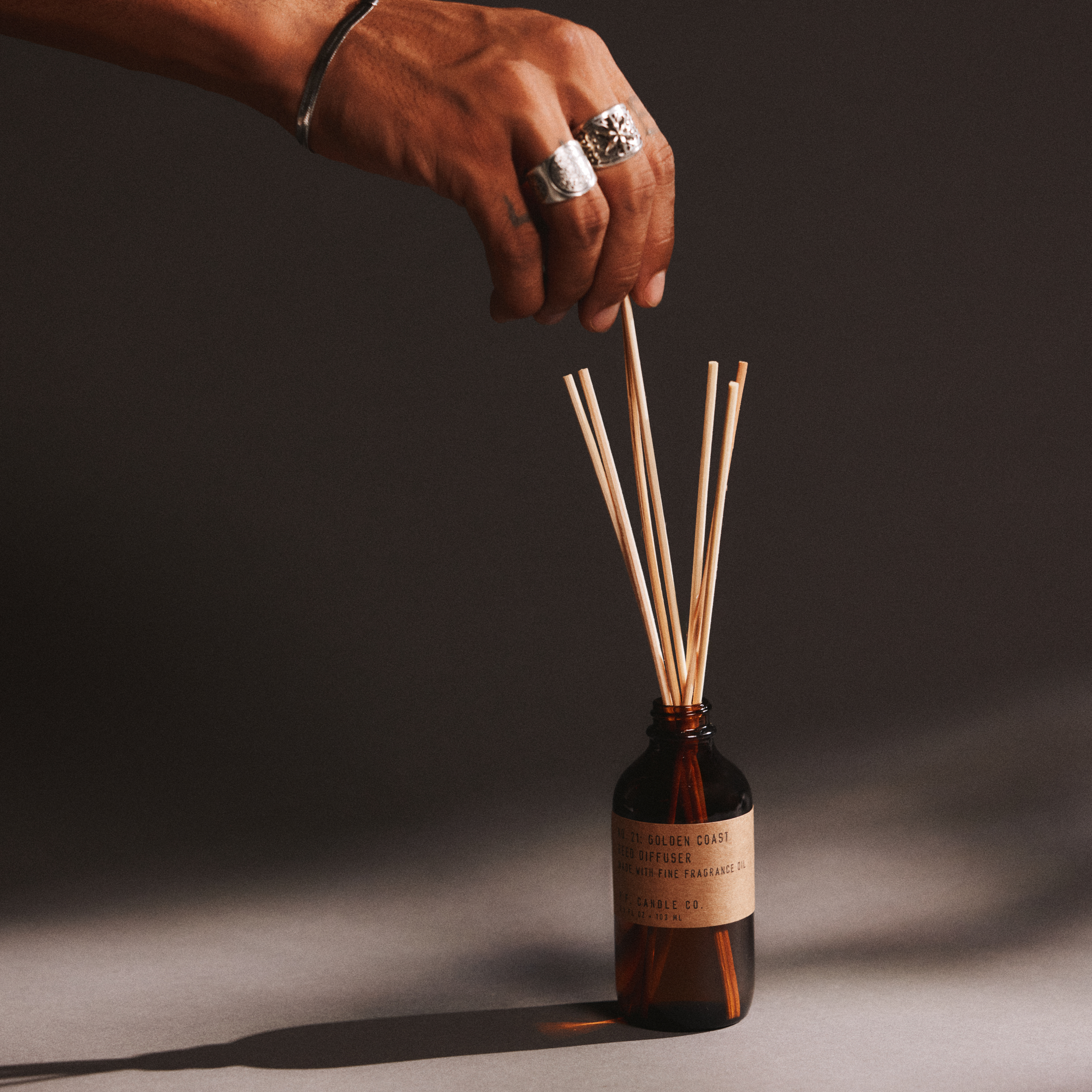 Hand with a ring holding reed diffuser sticks next to a bottle of golden coast reed diffuser on a dark background