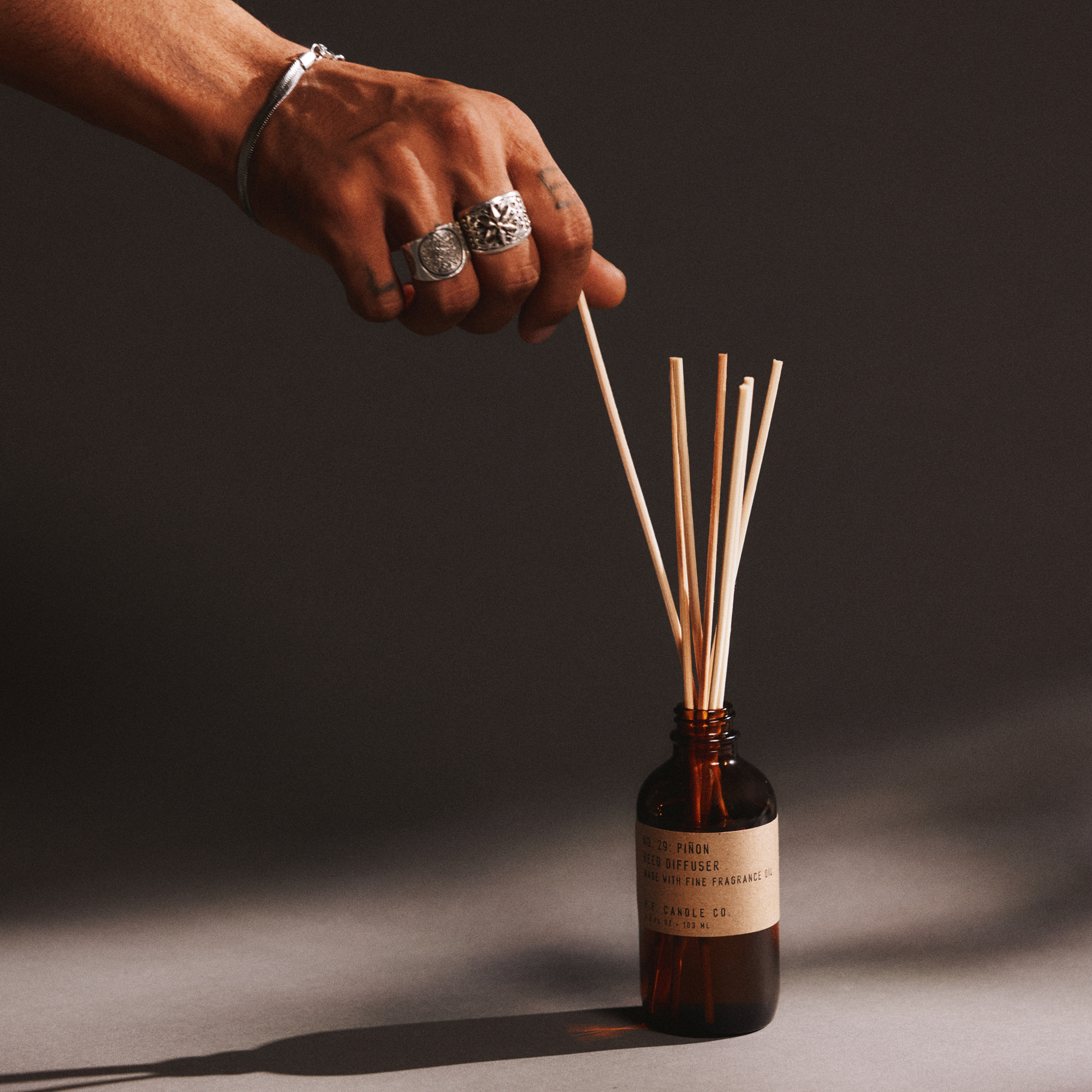 Hand with a ring holding reed diffuser sticks next to a bottle of pinon reed diffuser on a dark background