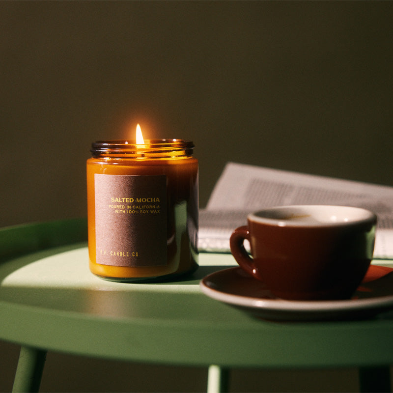 pf candle co Salted Mocha standard soy candle lit on top of a green table next the a newspaper and coffee mug