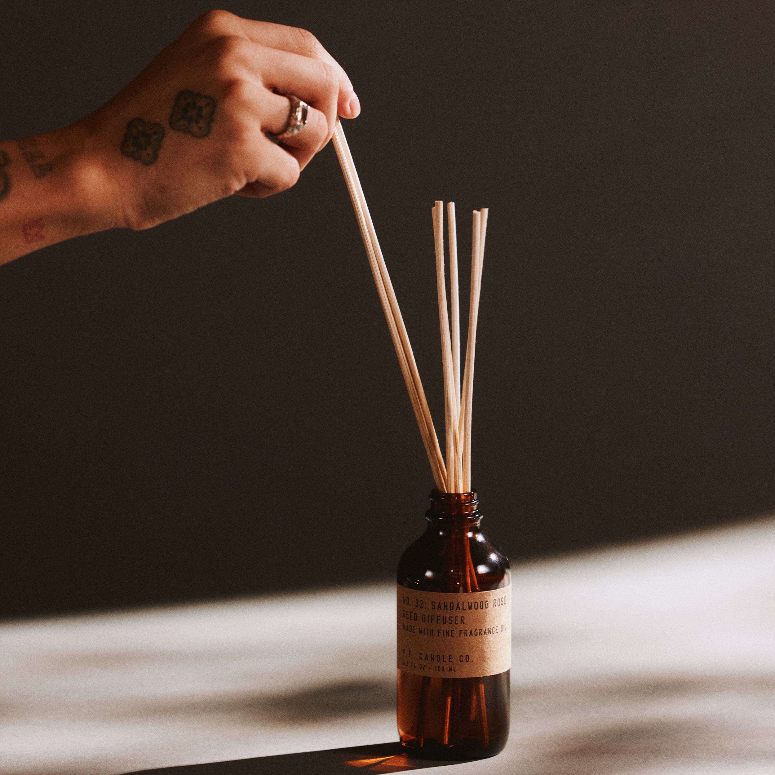 Hand with a ring holding reed diffuser sticks next to a bottle of sandalwood rose reed diffuser on a dark background
