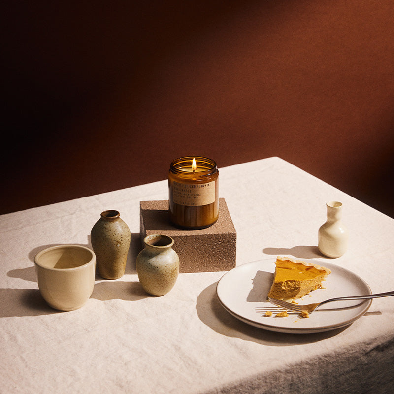 Lit pf candle co no. 1 Spiced Pumpkin amber jar candle displayed on a pedestal alongside ceramic vases and a cup, with a slice of pumpkin pie on a white plate in warm autumn lighting.
