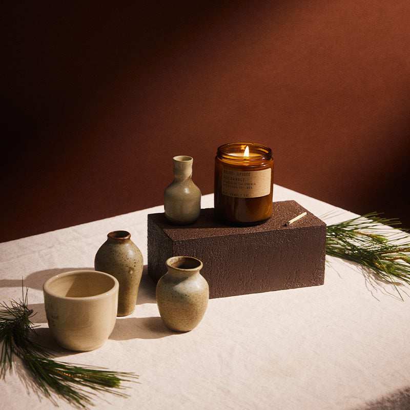 Lit pf candle co no. 5 Spruce amber jar candle displayed on a pedestal alongside ceramic vases and a cup, spruce pine leafs on a white plate in warm autumn lighting.