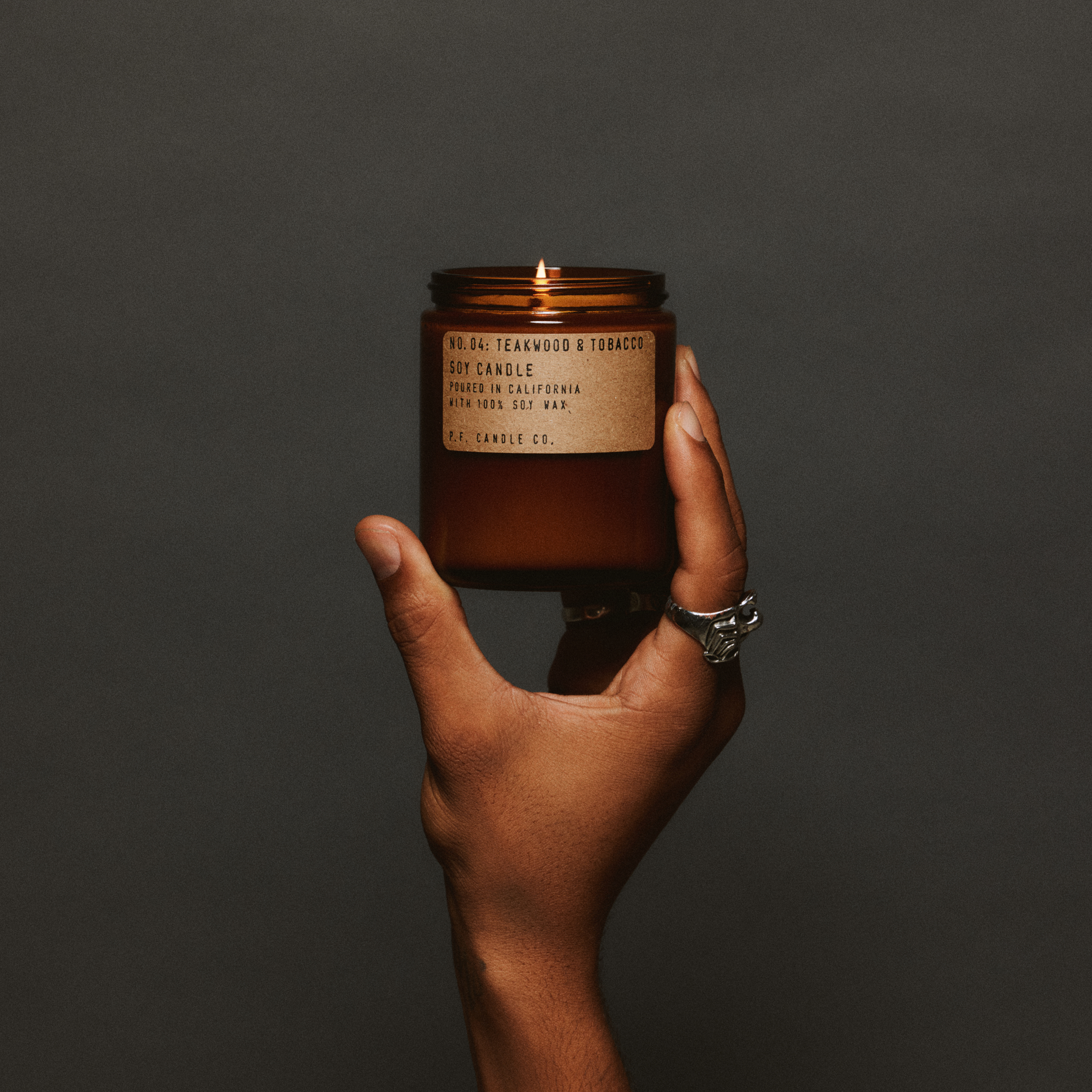 Hand holding a lit teakwood & tobacco standard candle with a label against a dark background