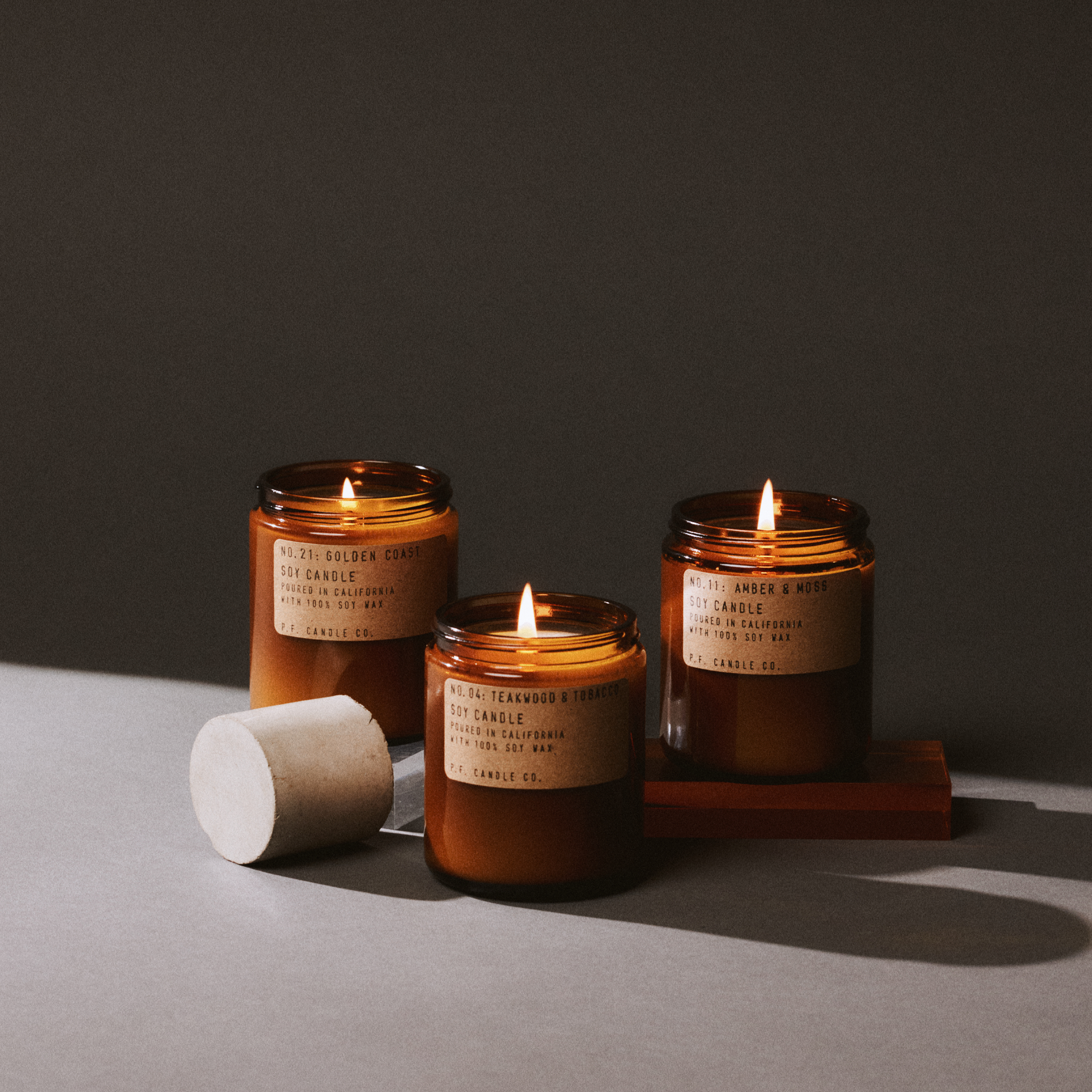 Three lit candles in amber jars with wooden lids on a gray surface.