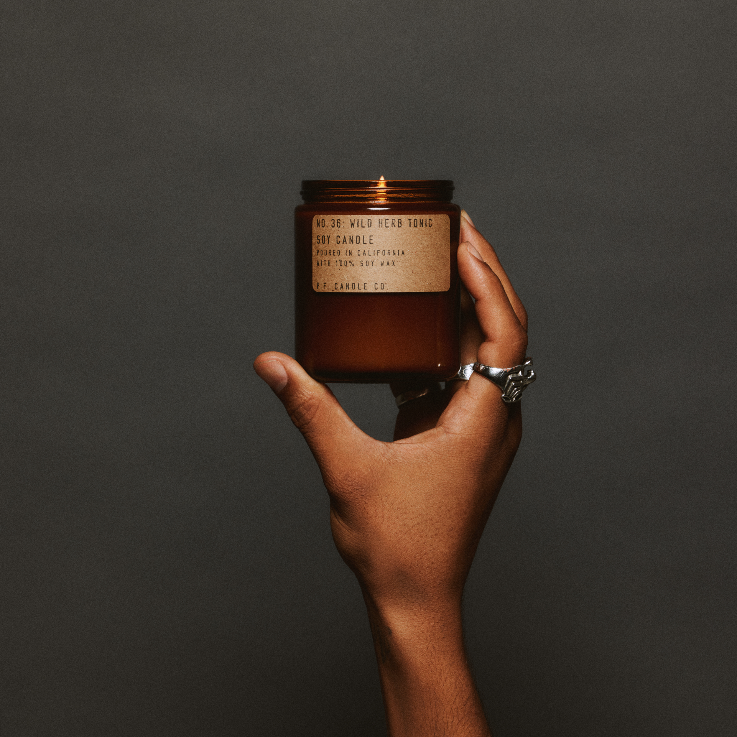 Hand holding a lit wild herb tonic standard candle with a label against a dark background